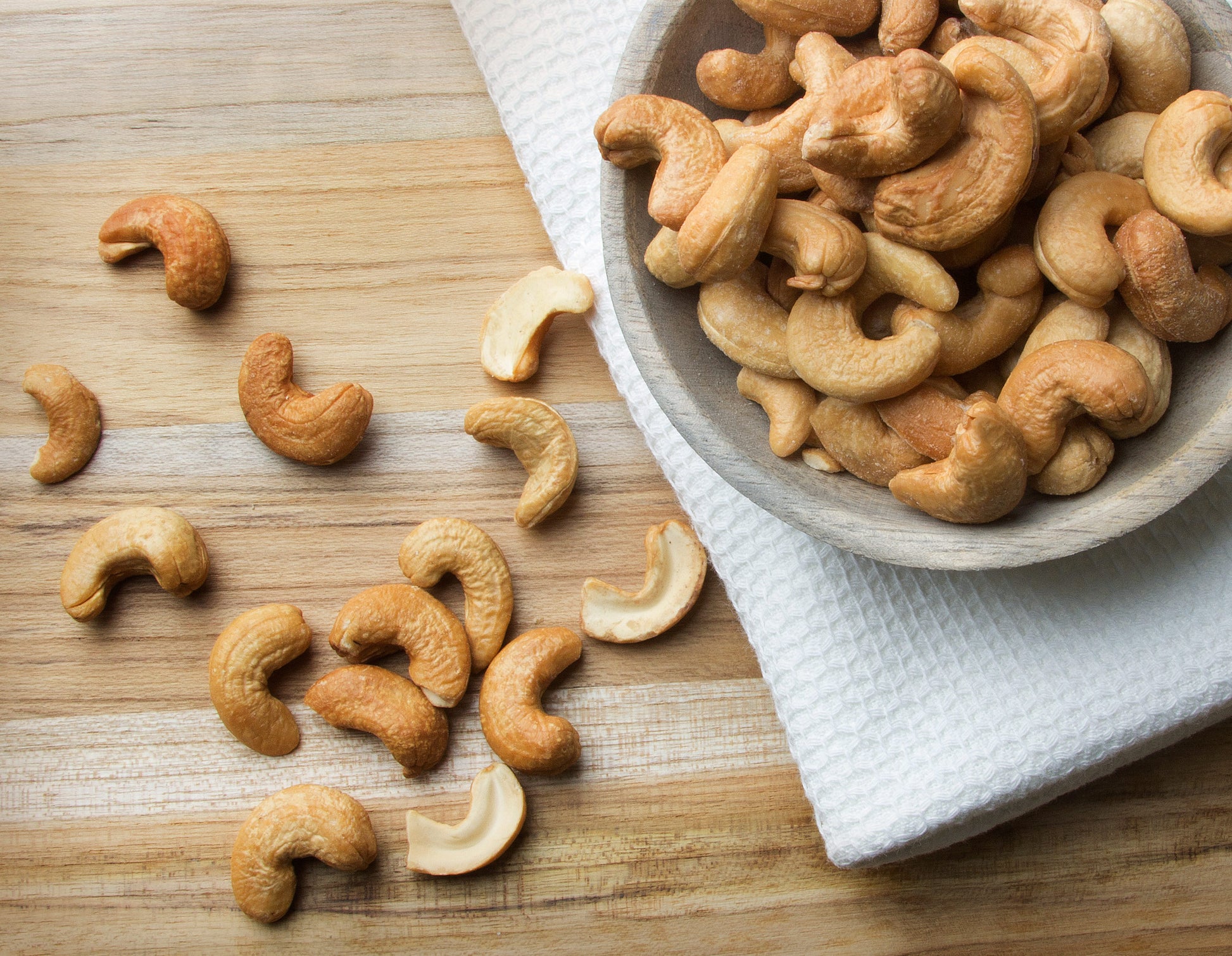 Cashews on a wooden surface with a bowl of cashews nearby.